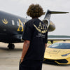 Person wearing a black t-shirt with gold lettering in front of a black airplane and gold car.