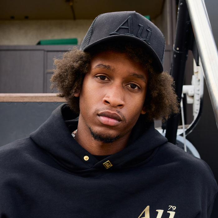 Man wearing a black hoodie and cap with visible "Au" branding, standing outdoors.