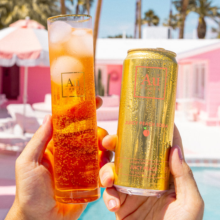 Two hands holding a glass of orange juice and a can of Au Peach Nectar by a poolside.