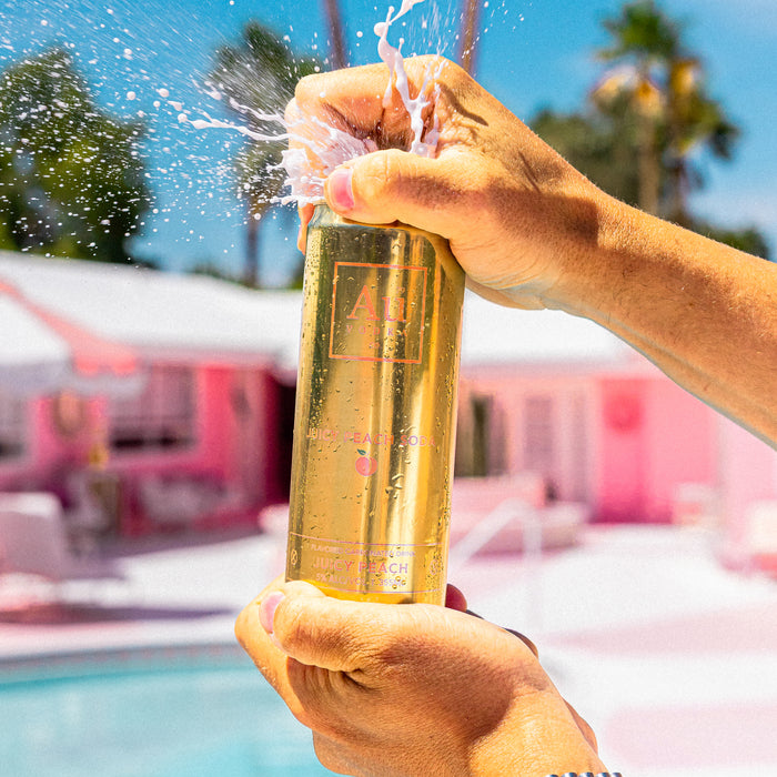 Person opening a gold Au Vodka can by a pool with a pink house and palm trees in the background