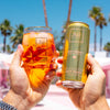 Two hands holding a glass of orange juice and a can of AuJuicy Peach against a palm tree background.