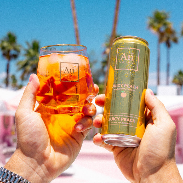 Two hands holding a glass of orange juice and a can of AuJuicy Peach against a palm tree background.