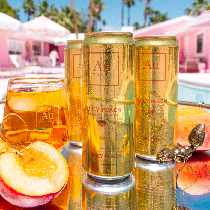 Cans of Au Peach sparkling water with a glass of the drink and peaches on a reflective surface.