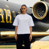 Person wearing a white t-shirt with gold logo in front of a black and gold airplane