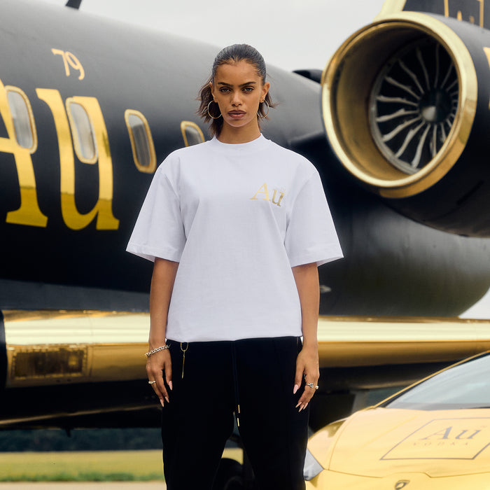 Person wearing a white t-shirt with gold logo in front of a black and gold airplane