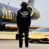 Person wearing a black hoodie with 'Au' logo in front of a black and gold airplane and golf sports car.
