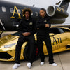 Two people in black hoodies with 'Au' branding in front of a gold car and black airplane.