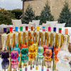 Colorful bottles with gold accents on a reflective surface outdoors with trees and a building in the background.
