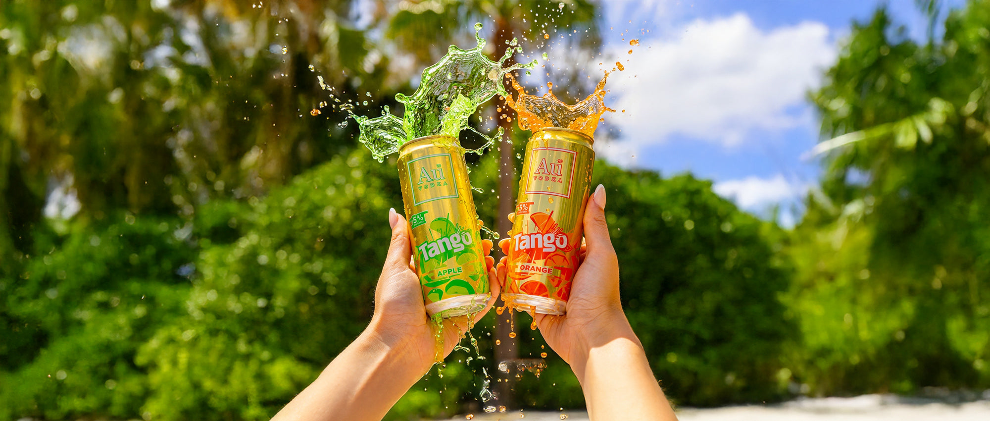 Two hands holding Au Vodka Tango Apple and Orange cans with liquid splashing out, tropical background
