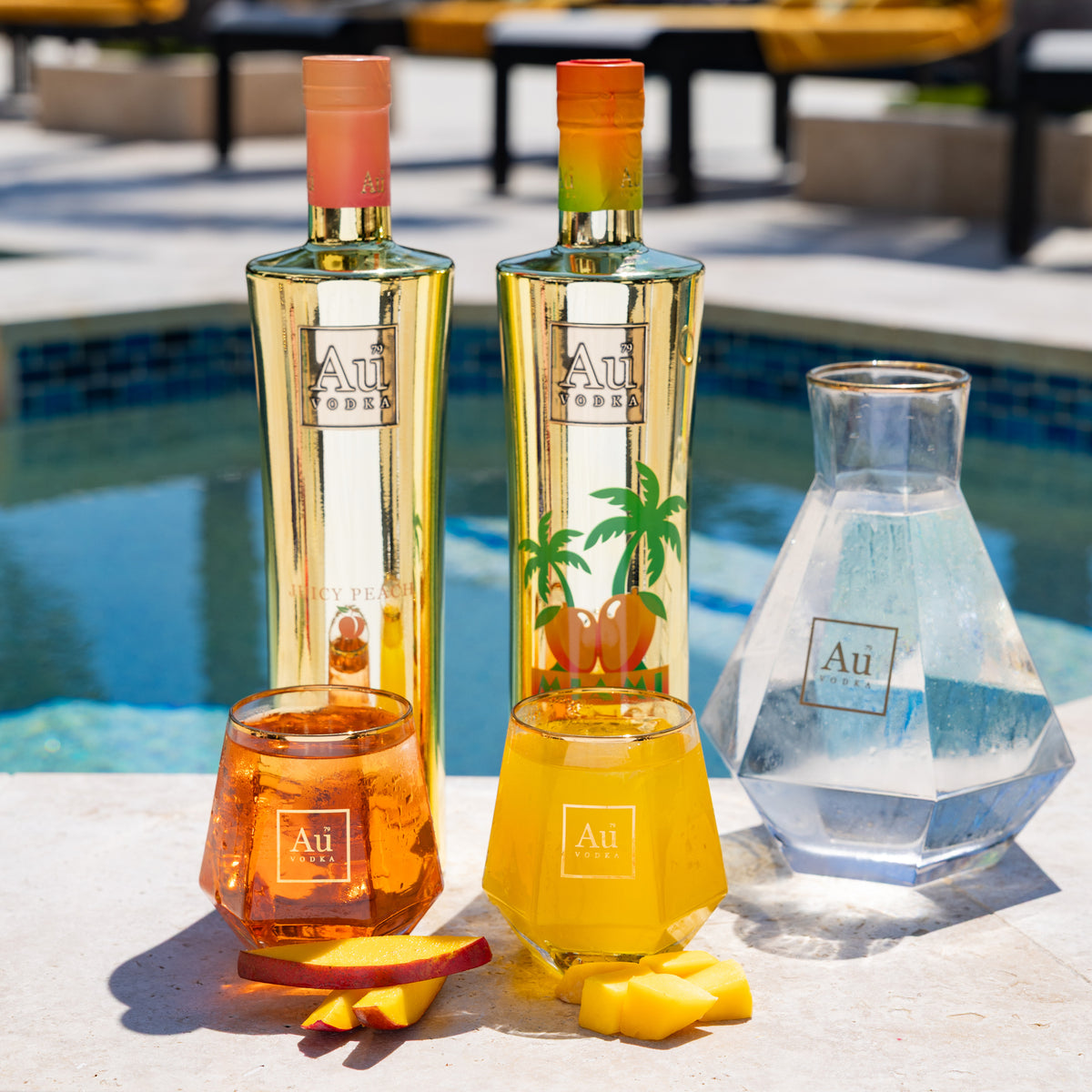 Two Au Vodka bottles, two glasses of cocktails, and a water carafe by a pool