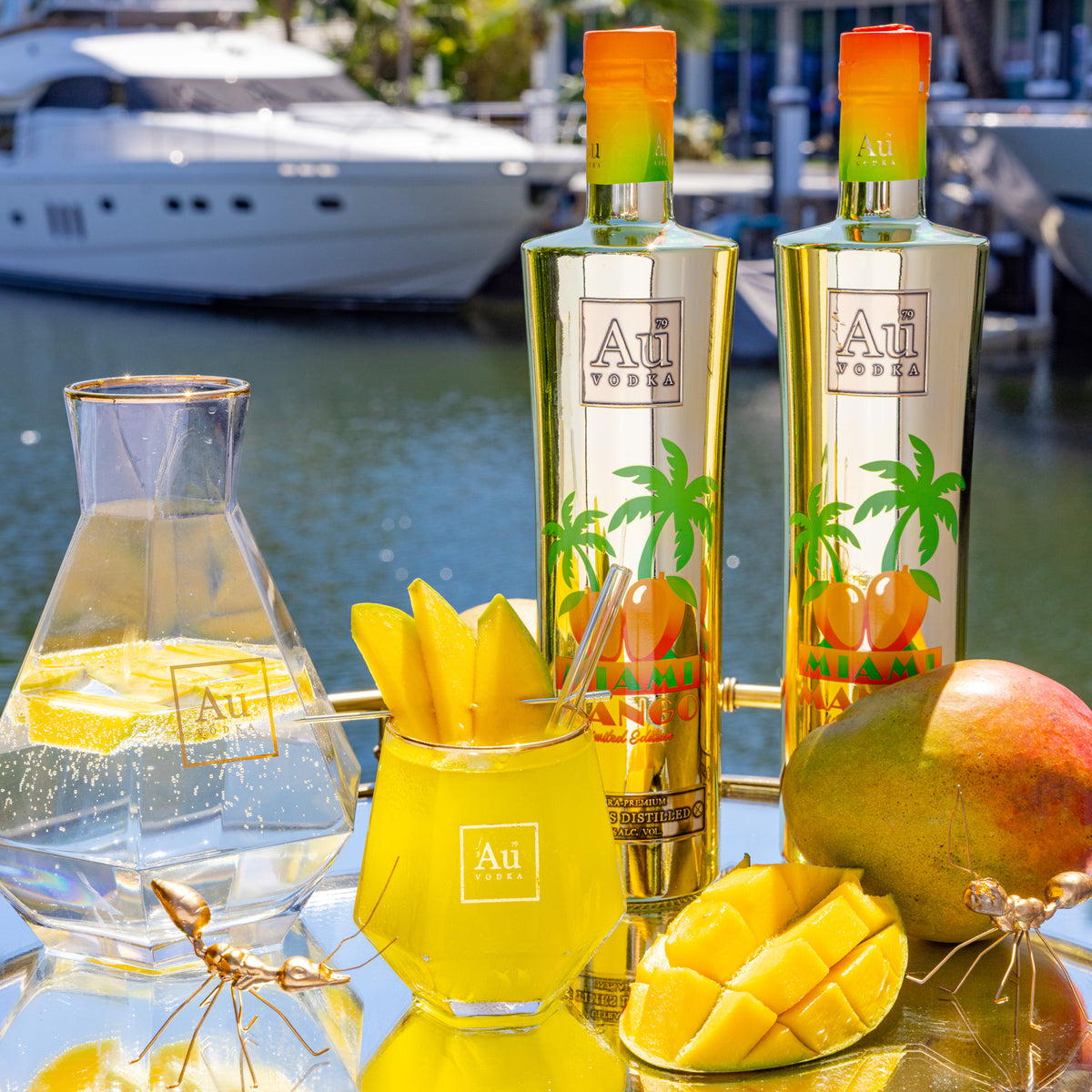 Au Vodka Miami Mango bottles, cocktail, and fresh mango on a mirrored tray with a yacht in background