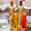 Two bottles of Au Vodka with two glasses filled with orange and red drinks on a snowy background.