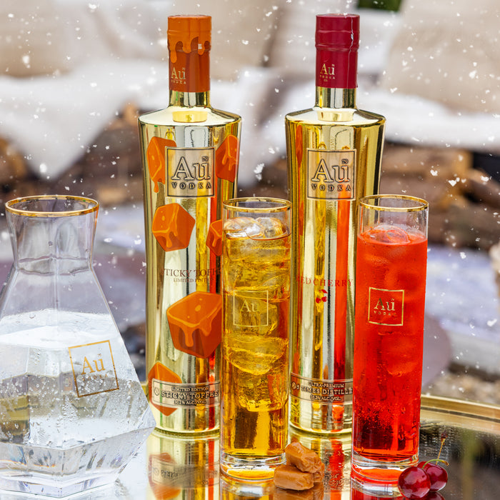 Two bottles of Au Vodka with two glasses filled with orange and red drinks on a snowy background.
