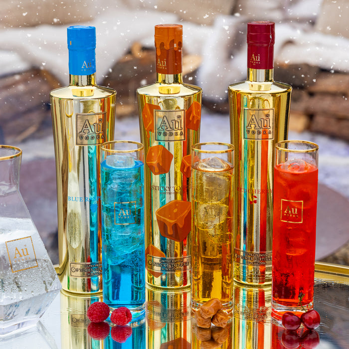 Three bottles of Au Vodka with corresponding colored glasses on a reflective surface.