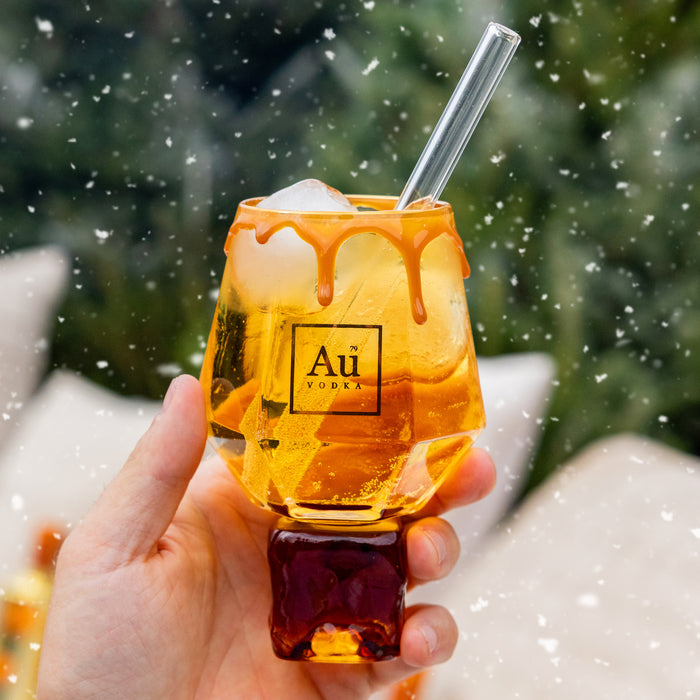 Hand holding a glass of orange liquid with a straw, against a blurred green and white background.
