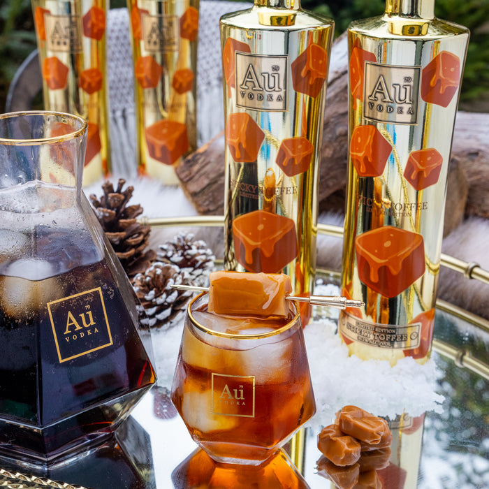 Au Vodka bottles with caramel candies and a glass of iced coffee on a reflective surface.