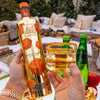 Person holding a bottle of Au Vodka Sticky Toffee and a glass with Appletiser, outdoor setting with Christmas decorations.