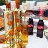 Two bottles of Au Vodka being poured into a glass with Coca-Cola bottles in the background.