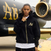 Person wearing a black hoodie with gold 'Au' lettering in front of jet.